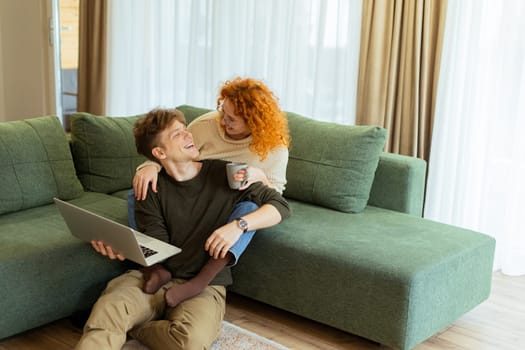 Two friends engage in a light-hearted moment at home, seated comfortably on a green couch, as they collaborate on a laptop, filled with laughter and joy.
