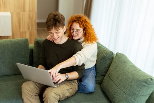 In a stylish living room, a couple shares a warm moment as they sit on a green couch, working on a laptop. The sunlight bathes them, perfect for a relaxed afternoon