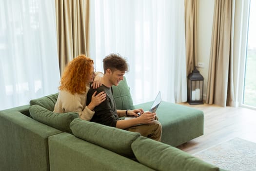 Two friends engage in a light-hearted moment at home, seated comfortably on a green couch, as they collaborate on a laptop, filled with laughter and joy.
