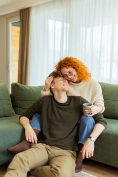 A couple enjoys a relaxing afternoon at home, seated comfortably on a plush sofa, exchanging loving smiles and laughter while sharing a warm beverage together