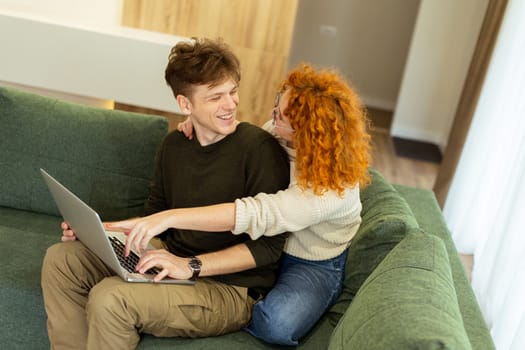 Two friends engage in a light-hearted moment at home, seated comfortably on a green couch, as they collaborate on a laptop, filled with laughter and joy.