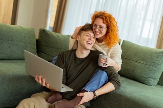 Two friends engage in a light-hearted moment at home, seated comfortably on a green couch, as they collaborate on a laptop, filled with laughter and joy.