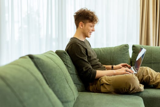 Enjoying a moment of productivity, a young individual sits on a comfortable green sofa, typing intently on a laptop, surrounded by bright, airy curtains that let in sunlight.