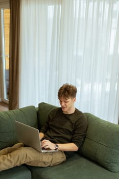 Focused on his laptop, a young man sits comfortably on a lush green couch, surrounded by light flooding through sheer curtains in a modern living space