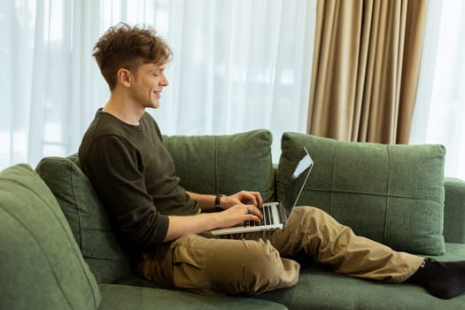 A young man sits comfortably on a green sofa, deeply focused on his laptop. Sunlight pours through large windows, creating a warm atmosphere in the inviting space.