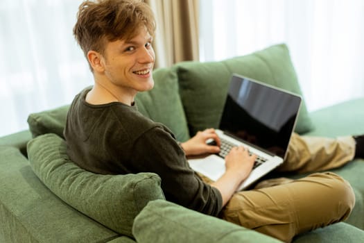A young man sits comfortably on a green sofa, deeply focused on his laptop. Sunlight pours through large windows, creating a warm atmosphere in the inviting space.