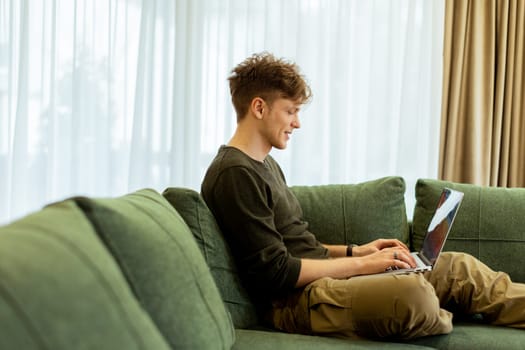A young man sits comfortably on a green sofa, deeply focused on his laptop. Sunlight pours through large windows, creating a warm atmosphere in the inviting space.