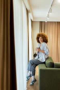 A young individual with curly hair sits on a green sofa, holding a warm cup of coffee, smiling while enjoying the serene ambiance of a stylish living space in soft morning light