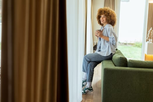 A young individual with curly hair sits on a green sofa, holding a warm cup of coffee, smiling while enjoying the serene ambiance of a stylish living space in soft morning light