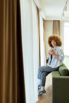 A young individual with curly hair sits on a green sofa, holding a warm cup of coffee, smiling while enjoying the serene ambiance of a stylish living space in soft morning light