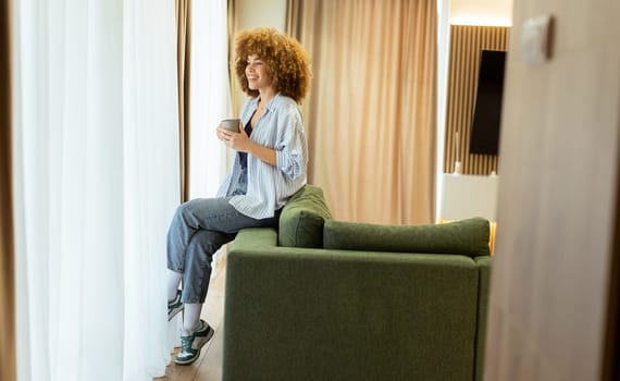 A young individual with curly hair sits on a green sofa, holding a warm cup of coffee, smiling while enjoying the serene ambiance of a stylish living space in soft morning light