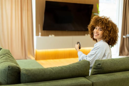 A woman with curly hair beams with joy as she sits on a soft couch, ready to enjoy her favorite show while holding the remote control in a modern living space