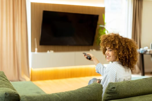 A woman with curly hair beams with joy as she sits on a soft couch, ready to enjoy her favorite show while holding the remote control in a modern living space