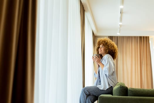 A young woman with curly hair is seated on a soft sofa, holding a warm drink and looking cheerfully towards the camera, with sunlight streaming through the window