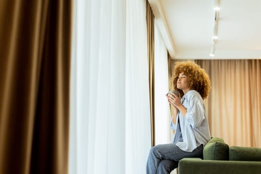 A young woman with curly hair is seated on a soft sofa, holding a warm drink and looking cheerfully towards the camera, with sunlight streaming through the window