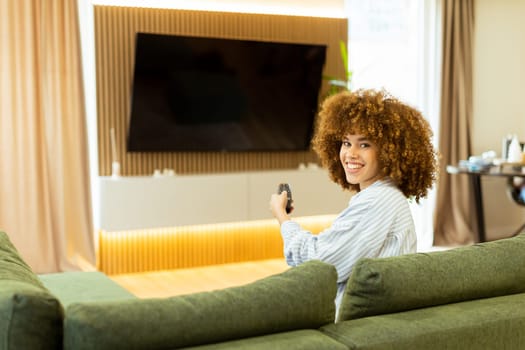 A woman with curly hair beams with joy as she sits on a soft couch, ready to enjoy her favorite show while holding the remote control in a modern living space