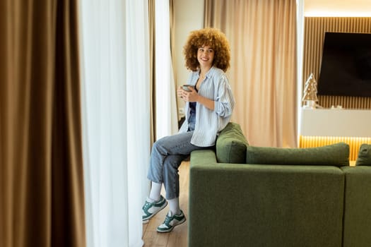 A young individual with curly hair sits on a green sofa, holding a warm cup of coffee, smiling while enjoying the serene ambiance of a stylish living space in soft morning light