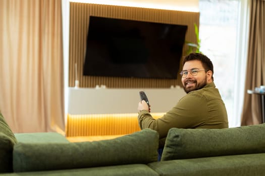 A man sits comfortably on a green couch, holding a remote, engaged in his favorite show. Soft lighting and modern decor create a serene ambiance perfect for unwinding.