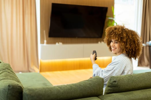 A woman with curly hair beams with joy as she sits on a soft couch, ready to enjoy her favorite show while holding the remote control in a modern living space