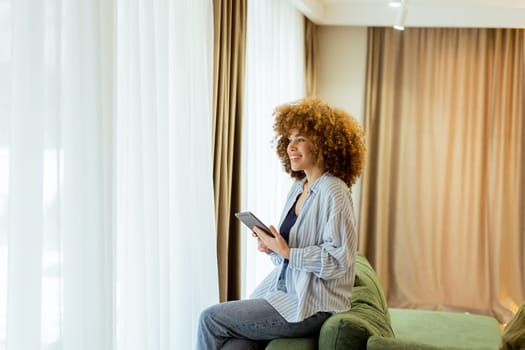 A woman with curly hair enjoys using her tablet, seated comfortably on a couch in a bright, inviting living space, surrounded by soft curtains and warm lighting