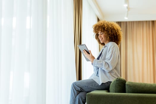 A woman with curly hair enjoys using her tablet, seated comfortably on a couch in a bright, inviting living space, surrounded by soft curtains and warm lighting