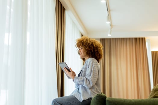 A woman with curly hair enjoys using her tablet, seated comfortably on a couch in a bright, inviting living space, surrounded by soft curtains and warm lighting