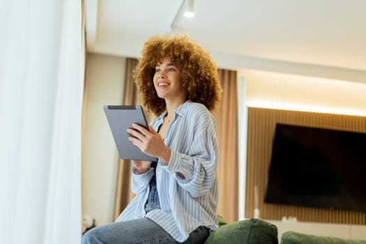 A young person with curly hair sits comfortably on a stylish couch, focused on their tablet. Warm light filters through sheer curtains, creating a cozy and inviting atmosphere.