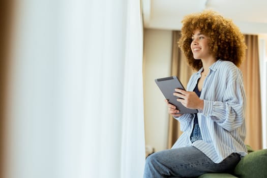 A young person with curly hair sits comfortably on a stylish couch, focused on their tablet. Warm light filters through sheer curtains, creating a cozy and inviting atmosphere.
