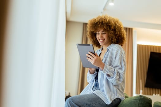 A young person with curly hair sits comfortably on a stylish couch, focused on their tablet. Warm light filters through sheer curtains, creating a cozy and inviting atmosphere.