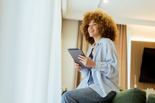 A young person with curly hair sits comfortably on a stylish couch, focused on their tablet. Warm light filters through sheer curtains, creating a cozy and inviting atmosphere.