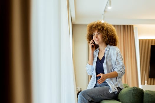 A joyful woman with curly hair chats on her phone, sitting comfortably on a green couch in a stylish room. Warm sunlight filters through the curtains, creating an inviting atmosphere