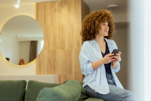 A woman with curly hair sits comfortably on a green sofa, smiling as she interacts with her smartphone in a bright and inviting living room adorned with contemporary design