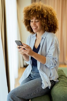 With a bright smile, a young woman sits on a green sofa, engaged with her smartphone amid a warm and inviting indoor space filled with natural light.