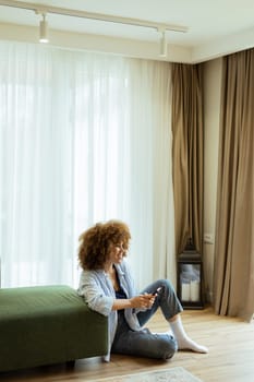 Comfort and style combine in a serene living space, featuring a woman with curly hair enjoying her time on a smartphone by a green sofa. Soft sunlight fills the airy room
