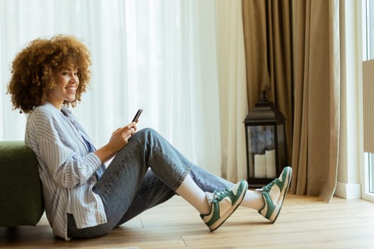 A young woman with curly hair sits on the wooden floor, smiling while looking at her smartphone in a sunlit room. Soft curtains sway gently as she enjoys her peaceful moment