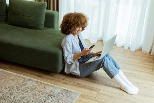 Seated comfortably on the floor, a young woman engages with her laptop while checking her smartphone. The cozy, sunlit living room creates a serene backdrop for her dual focus