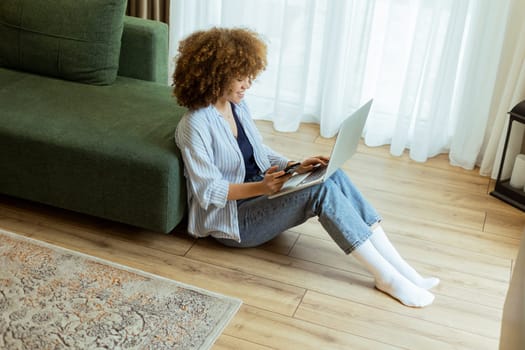 Seated comfortably on the floor, a young woman engages with her laptop while checking her smartphone. The cozy, sunlit living room creates a serene backdrop for her dual focus