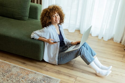 Seated comfortably on the floor, a young woman engages with her laptop while checking her smartphone. The cozy, sunlit living room creates a serene backdrop for her dual focus