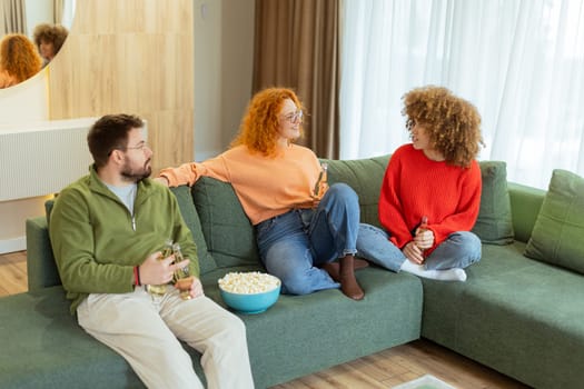 Three friends gather in a cozy living room, sharing drinks and popcorn while chatting and laughing. The warm atmosphere invites relaxation and connection among them.