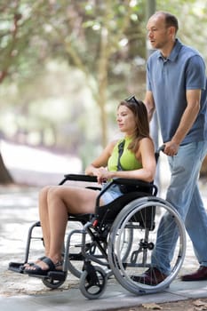 Man assisting a disabled woman in a wheelchair, enjoying a stroll through a peaceful park