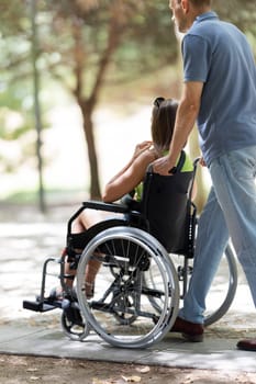 Man assisting a disabled woman in a wheelchair, enjoying a walk through a sunny park
