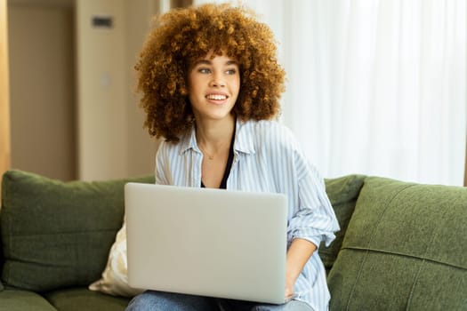 Sitting comfortably on a plush green sofa, a young woman types on her laptop, basking in the warm sunlight streaming through the large windows, radiating focus and tranquility.