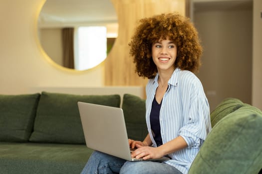 Sitting comfortably on a plush green sofa, a young woman types on her laptop, basking in the warm sunlight streaming through the large windows, radiating focus and tranquility.