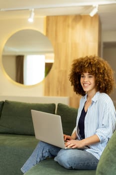 Sitting comfortably on a plush green sofa, a young woman types on her laptop, basking in the warm sunlight streaming through the large windows, radiating focus and tranquility.