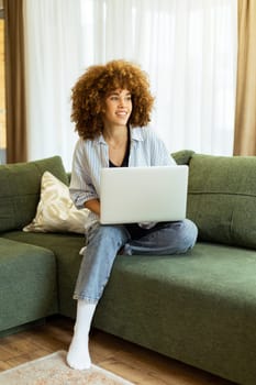 Sitting comfortably on a plush green sofa, a young woman types on her laptop, basking in the warm sunlight streaming through the large windows, radiating focus and tranquility.