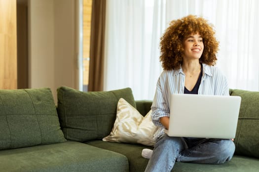 Sitting comfortably on a plush green sofa, a young woman types on her laptop, basking in the warm sunlight streaming through the large windows, radiating focus and tranquility.
