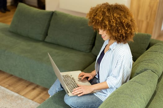 Sitting comfortably on a plush green sofa, a young woman types on her laptop, basking in the warm sunlight streaming through the large windows, radiating focus and tranquility.