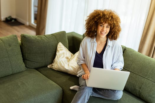 Sitting comfortably on a plush green sofa, a young woman types on her laptop, basking in the warm sunlight streaming through the large windows, radiating focus and tranquility.