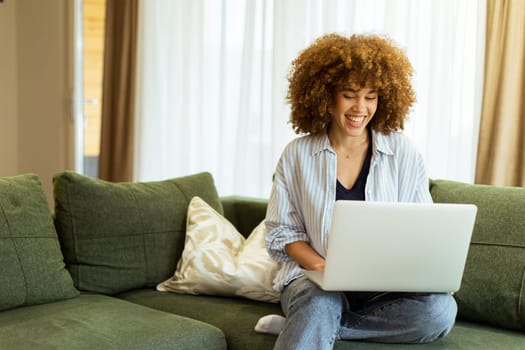 Sitting comfortably on a plush green sofa, a young woman types on her laptop, basking in the warm sunlight streaming through the large windows, radiating focus and tranquility.
