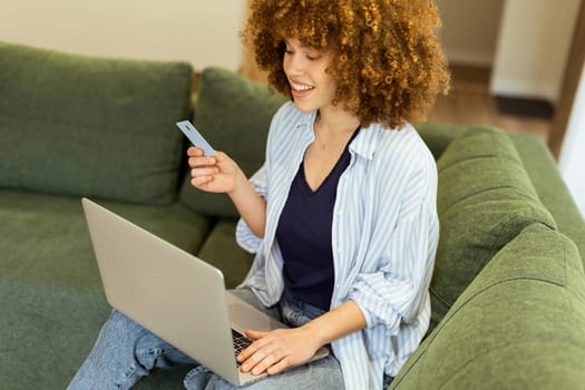 A young woman with curly hair happily shops online, holding a credit card, while seated on a comfortable couch in a stylish living room filled with warm sunlight.